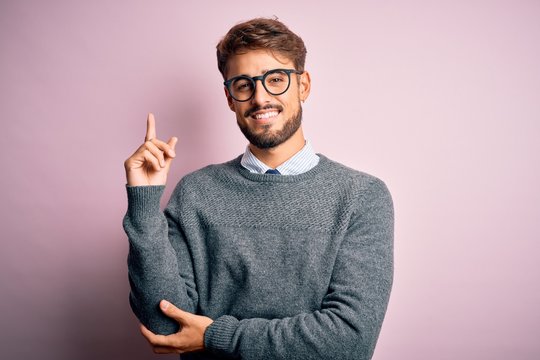 Young handsome man with beard wearing glasses and sweater standing over pink background with a big smile on face, pointing with hand and finger to the side looking at the camera.