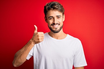 Young handsome man with beard wearing casual t-shirt standing over red background doing happy thumbs up gesture with hand. Approving expression looking at the camera showing success.