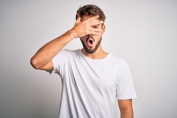 Young handsome man with beard wearing casual t-shirt standing over white background peeking in shock covering face and eyes with hand, looking through fingers with embarrassed expression.