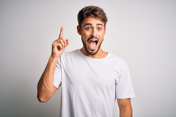 Fototapeta premium Young handsome man with beard wearing casual t-shirt standing over white background pointing finger up with successful idea. Exited and happy. Number one.