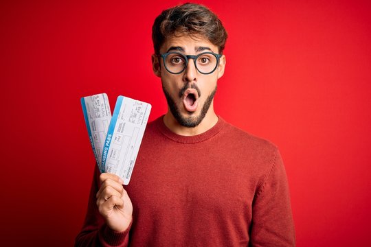 Young tourist man on vacation holding boarding pass standing over red bakground scared in shock with a surprise face, afraid and excited with fear expression