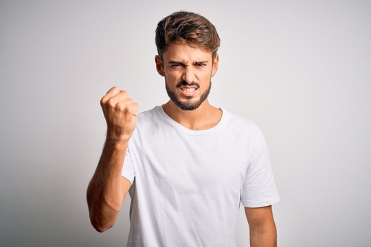 Young handsome man with beard wearing casual t-shirt standing over white background angry and mad raising fist frustrated and furious while shouting with anger. Rage and aggressive concept.