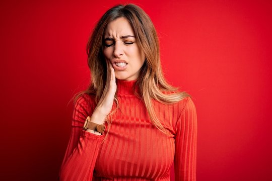 Young beautiful brunette woman wearing casual turtleneck sweater over red background touching mouth with hand with painful expression because of toothache or dental illness on teeth. Dentist