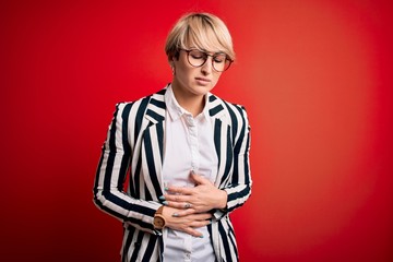 Blonde business woman with short hair wearing glasses and striped jacket over red background with hand on stomach because indigestion, painful illness feeling unwell. Ache concept.
