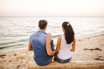 Theme family weekend, beach summer vacation with child. Young stylish family of three dad mom and daughter sit one year with their backs on lying tree, log overlooking horizon sea sunset in summer