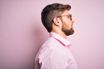 Young handsome blond man with beard and blue eyes wearing pink shirt and glasses looking to side,...