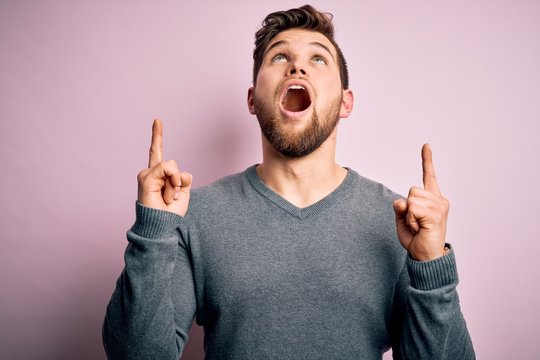 Young handsome blond man with beard and blue eyes wearing casual sweater amazed and surprised looking up and pointing with fingers and raised arms.
