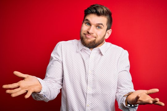 Young blond businessman with beard and blue eyes wearing elegant shirt over red background clueless and confused expression with arms and hands raised. Doubt concept.