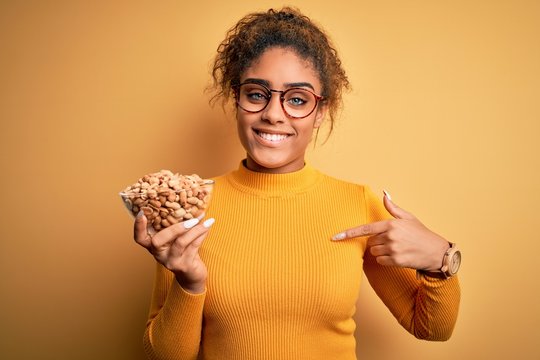 Young african american afro girl holding bowl with healthy peanuts over yellow background with surprise face pointing finger to himself