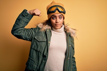 Young african american afro skier girl wearing snow sportswear and ski goggles Strong person showing arm muscle, confident and proud of power
