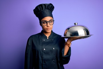 Young african american cooker girl wearing uniform and hat holding tray with dome with a confident expression on smart face thinking serious