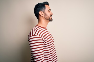 Young handsome man with beard wearing casual striped t-shirt standing over white background looking to side, relax profile pose with natural face with confident smile.