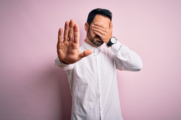 Young handsome man with beard wearing casual shirt standing over pink background covering eyes with hands and doing stop gesture with sad and fear expression. Embarrassed and negative concept.