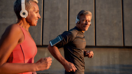 Sport making us happier. Positive and active middle-aged couple in headphones running through the city street in the morning