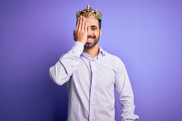 Young handsome man with beard wearing golden crown of king over purple background covering one eye with hand, confident smile on face and surprise emotion.