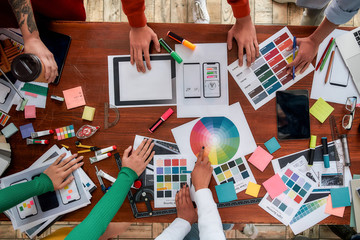 Mobile app design. Top view of designers discussing sketches, choosing colors from palettes lying on the desk while having a meeting in the modern office