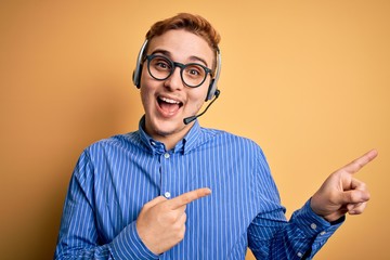 Young handsome redhead call center agent man wearing glasses working using headset smiling and looking at the camera pointing with two hands and fingers to the side.