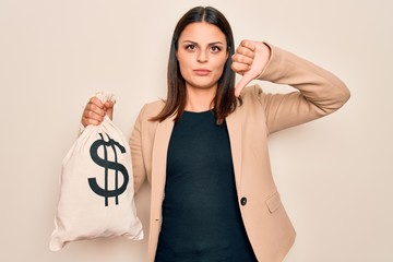 Young beautiful brunette woman holding bag with dollar symbol over white background with angry face, negative sign showing dislike with thumbs down, rejection concept