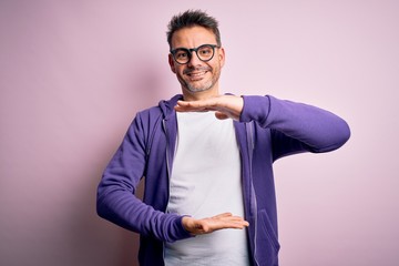 Young handsome man wearing purple sweatshirt and glasses standing over pink background gesturing with hands showing big and large size sign, measure symbol. Smiling looking at the camera. Measuring