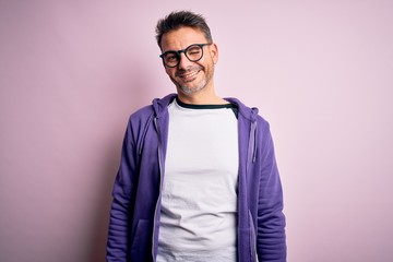 Young handsome man wearing purple sweatshirt and glasses standing over pink background winking...