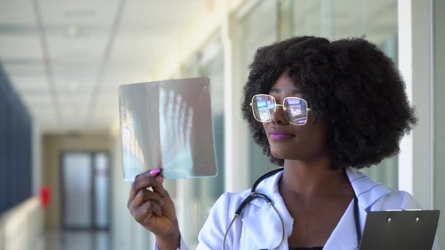 Female african-american doctor examines x-ray of leg, holding it in hands indoors. Specialist holds transparent image of leg in arms, and carefully researches it
