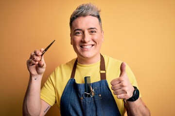 Young modern professional hairdresser man holding scissors over isolated yellow background happy with big smile doing ok sign, thumb up with fingers, excellent sign