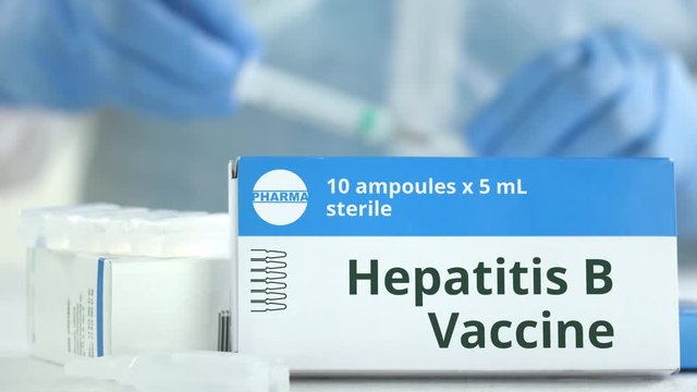Box with hepatitis B vaccine on the table against blurred lab assistant. Fictional phaceutical logo