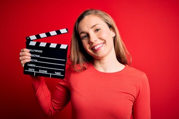 Young beautiful blonde clapper woman holding clapboard over isolated red background with a happy...
