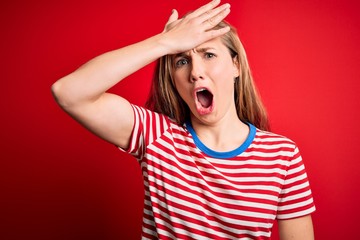 Young beautiful blonde woman wearing casual striped t-shirt over isolated red background surprised with hand on head for mistake, remember error. Forgot, bad memory concept.