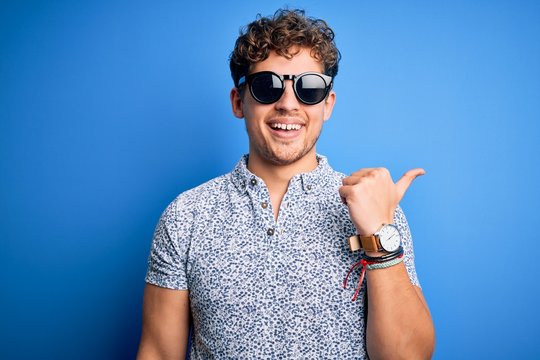 Young blond handsome man with curly hair on vacation wearing striped polo and sunglasses smiling with happy face looking and pointing to the side with thumb up.