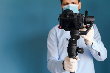 Front view on caucasian adult man blogger standing in front of the blue wall background holding gimbal stabilizer with camera making video about pandemic virus disease spread wearing protective mask