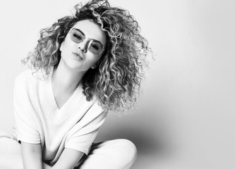 Black and white photo, blonde curly lady in sunglasses, suit of pants and blouse. Kissing you, sitting on floor isolated on white