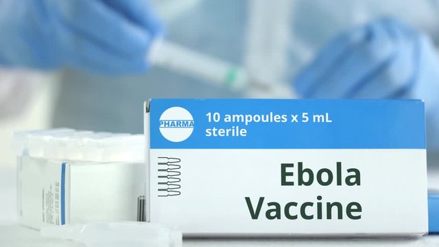 Ampoules with ebola vaccine on the table near working laboratory assistant, fictional logo on the box