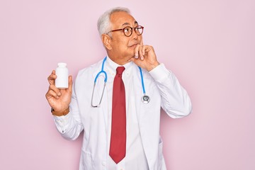 Middle age senior grey-haired doctor man holding prescription pills over pink background serious face thinking about question, very confused idea