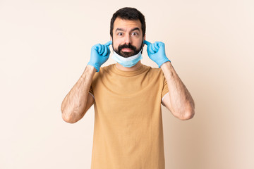 Caucasian man with beard protecting from the coronavirus with a mask and gloves over isolated background frustrated and covering ears