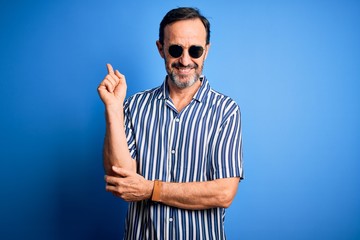 Naklejka premium Middle age hoary man wearing striped shirt and sunglasses over isolated blue background with a big smile on face, pointing with hand and finger to the side looking at the camera.