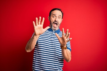 Naklejka premium Middle age hoary man wearing casual striped polo standing over isolated red background afraid and terrified with fear expression stop gesture with hands, shouting in shock. Panic concept.