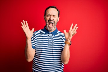 Fototapeta premium Middle age hoary man wearing casual striped polo standing over isolated red background celebrating mad and crazy for success with arms raised and closed eyes screaming excited. Winner concept