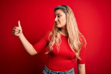 Young beautiful blonde woman wearing casual t-shirt standing over isolated red background Looking proud, smiling doing thumbs up gesture to the side