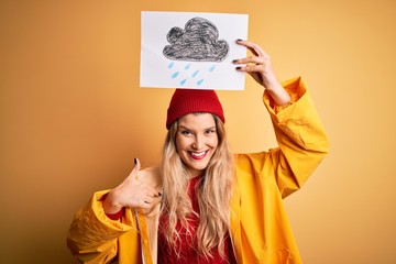 Young beautiful blonde woman wearing raincoat and wool cap holding banner with cloud image with surprise face pointing finger to himself