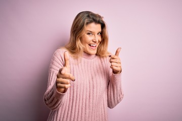 Young beautiful blonde woman wearing casual pink sweater over isolated background pointing fingers to camera with happy and funny face. Good energy and vibes.