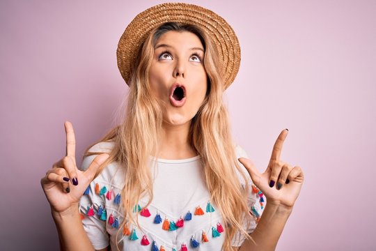 Young beautiful blonde woman wearing t-shirt and hat over isolated pink background amazed and surprised looking up and pointing with fingers and raised arms.