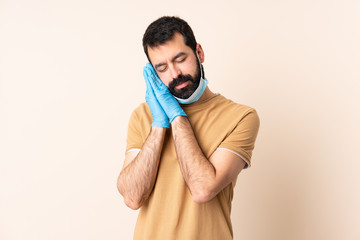 Caucasian man with beard protecting from the coronavirus with a mask and gloves over isolated background making sleep gesture in dorable expression