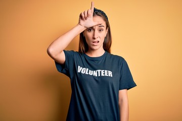 Young beautiful woman wearing volunteer t-shirt doing volunteering over yellow background making fun of people with fingers on forehead doing loser gesture mocking and insulting.