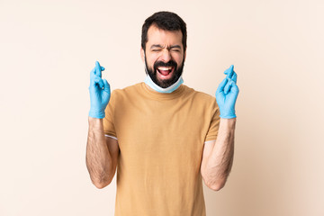 Caucasian man with beard protecting from the coronavirus with a mask and gloves over isolated background with fingers crossing