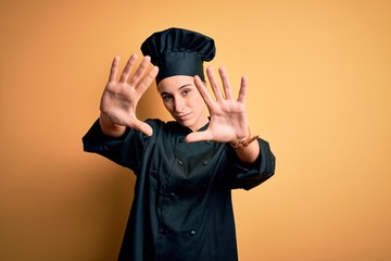 Young beautiful chef woman wearing cooker uniform and hat standing over yellow background doing frame using hands palms and fingers, camera perspective