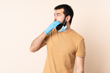 Caucasian man with beard protecting from the coronavirus with a mask and gloves over isolated background doing surprise gesture while looking to the side