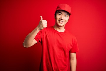 Young handsome chinese delivery man wearing cap standing over isolated red background doing happy thumbs up gesture with hand. Approving expression looking at the camera showing success.