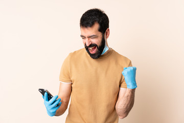 Caucasian man with beard protecting from the coronavirus with a mask and gloves over isolated background with phone in victory position