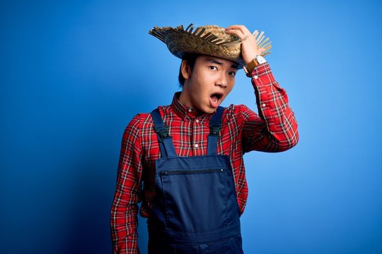 Young handsome chinese farmer man wearing apron and straw hat over blue background surprised with hand on head for mistake, remember error. Forgot, bad memory concept.